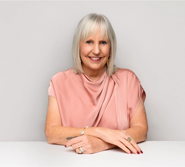 Older woman with straight gray hair, wearing a pink draped blouse, sits at a table smiling. She has rings, a bracelet, earrings, and neatly painted nails. The background is plain and light-colored. -Dooley Designs by Kim