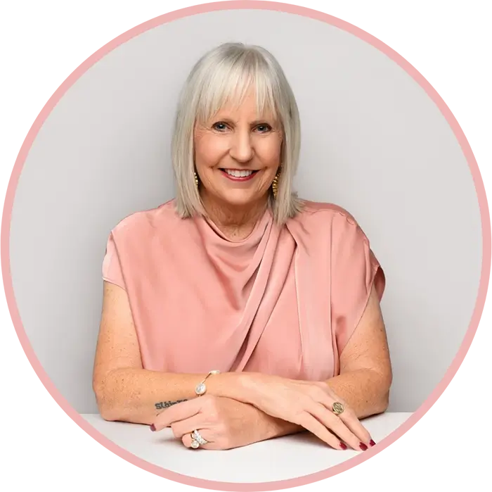 Smiling older woman with straight gray hair and bangs, wearing a pink blouse and jewelry, sits at a white table with her arms resting in front of her against a plain light background. Circular pink border frames the image. -Dooley Designs by Kim